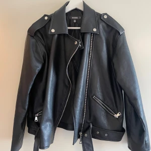 Svart skinnjacka från Missguided - Säljer en svart oversized skinnjacka från Missguided med silvriga dragkedjor, axelklaffar och bälte i midjan. Jackan har klassisk bikerstil och flera fickor med dragkedja. Sitter jätte fint på. Storlek 36. 