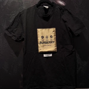 Svart t-shirt med beige Burberry-logga - Svart t-shirt från Burberry med en beige patch framtill där Burberry-loggan och text syns. Klassisk rund hals och korta ärmar. Perfekt för dig som gillar stilrena märkesplagg.