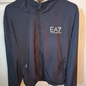Svart vindjacka från Emporio Armani EA7 - Säljer en svart vindjacka från Emporio Armani EA7 med huva och dragkedja framtill. Jackan har två fickor med dragkedja och en stilren logga på bröstet. Perfekt för dig som gillar sportig och clean stil. Bra skick men en reva på knappt en cm på ryggen. Digitalt kvitto finns.