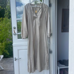Beige långklänning med v-ringning - Säljer en stilren beige långklänning med v-ringning och långa ärmar. Klänningen har en avslappnad passform och är tillverkad i ett glansigt material som ger en elegant känsla. Perfekt för sommarens tillfällen