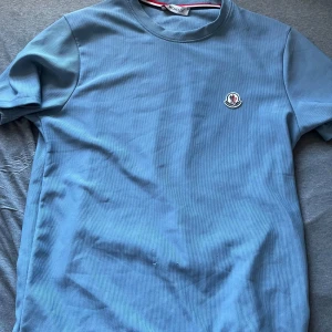 Blå t-shirt från Moncler - Säljer en stilren blå t-shirt från Moncler med rund hals och klassisk logga broderad på bröstet. T-shirten har korta ärmar och är tillverkad i ett mjukt bomullsmaterial. Perfekt för en clean och avslappnad look. Pris kan diskuteras 