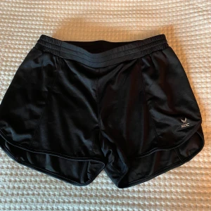 Svarta träningsshorts från SOC - Svarta träningsshorts från SOC med elastisk midja och sportig design. Lätta och luftiga med tekniskt material som håller dig torr och bekväm under träningen. Perfekta för gymmet eller löprundan.