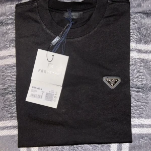 Svart t-shirt från Prada - Säljer en svart t-shirt från Prada med klassisk rund hals och diskret logotyp på bröstet. T-shirten är i mjuk bomull och har en stilren design som passar perfekt till många olika outfits.