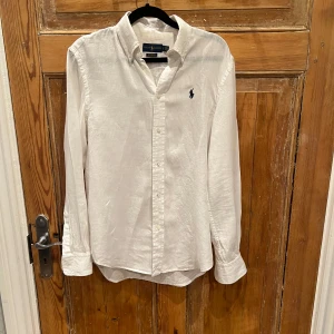Vit linneskjorta från Ralph Lauren - Klassisk vit linneskjorta från Ralph Lauren med relaxed fit. Skjortan har lång ärm, knappar framtill och den ikoniska broderade loggan på bröstet. Perfekt för en stilren och avslappnad look.