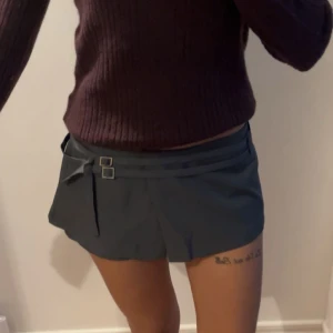 Mörkgrå kjol med bältdetalj  - Stilren mörkgrå kjol med två dekorativa bälten och spännen i midjan. Kjolen har en omlott-look framtill och dragkedja i sidan. Perfekta för en trendig och modern stil.