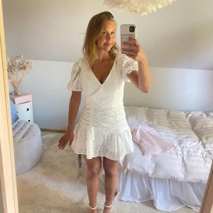 Vit kort klänning med puffärm och broderade detaljer - Superfin vit kortklänning med v-ringning, puffärmar och broderade hålmönster. Klänningen har omlottkänsla, rynkade detaljer framtill och knyt i midjan. Perfekt för sommarens alla tillfällen! Studentklänning student vit kort klänning