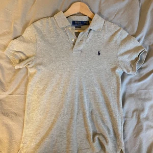 Ljusgrå pikétröja från Polo Ralph Lauren - Säljer en ljusgrå pikétröja från Polo Ralph Lauren med klassisk krage och två knappar. Slim fit-modell med korta ärmar och den ikoniska broderade loggan på bröstet. Perfekt för en stilren och avslappnad look.