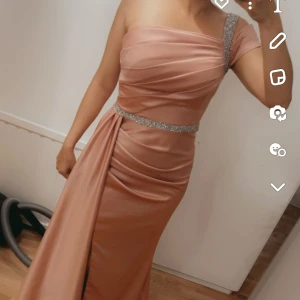 Champagnefärgad långklänning med glitterdetaljer - Säljer en elegant champagnefärgad långklänning med enaxlad design och glittriga detaljer vid axel och midja. Klänningen har draperad överdel och slits framtill för en snygg siluett. Perfekt för bal eller fest!