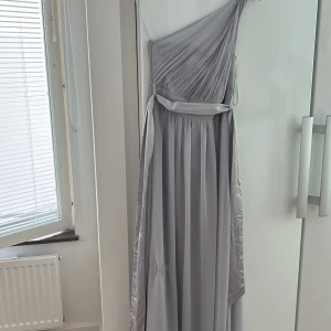 Ljusgrå långklänning med oneshoulder - Säljer en elegant ljusgrå långklänning med oneshoulder och draperad överdel. Klänningen har ett brett band i midjan och ett vackert fall. Perfekt för bal eller fest. Materialet känns mjukt och följsamt.