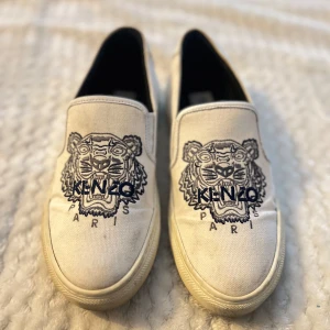Kenzo skor - Säljer ett par vita slip-on sneakers från Kenzo Paris med ikoniskt broderat tigermotiv och texten 'KENZO PARIS' framtill. Skorna har rund tå och är tillverkade i canvas med gummisula. Perfekta för dig som gillar streetstyle och vill sticka ut. Bara att höra av dig om du har några funderingar🌼💕