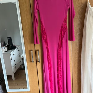 Rosa långklänning med spetsdetaljer asos  - Säljer en lång, figurnära rosa klänning med långa ärmar och dekorativa spetsinslag framtill och baktill. Klänningen har en elegant och modern look med insydda spetspaneler som ger extra detalj. Perfekt för dig som vill sticka ut.