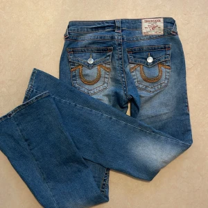 Blå True Religion jeans med broderade fickor - Säljer ett par blå jeans från True Religion med snygga broderade bakfickor och silverfärgade knappar. Jeansen har bootcut-modell och klassisk femficksdesign. 