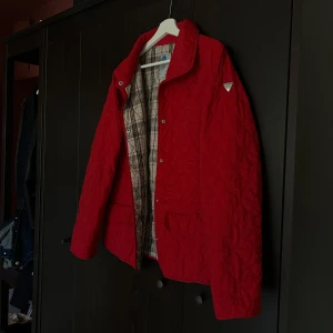 Röd quiltad underjacka i dun från Luhta - Snygg röd quiltad jacka från Luhta med rutigt innerfoder och tryckknappar framtill. Jackan har två stora fickor och en klassisk krage. Perfekt för dig som letar efter en riktig kvalitativ seglarjacka/ underjacka.