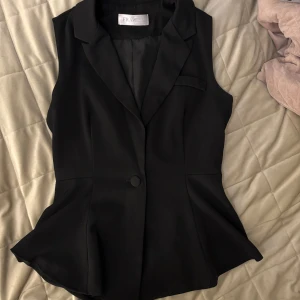 Svart kavajväst  - Stilren svart väst med kavajkrage och en knapp framtill. Västen har figurnära passform och lätt utsvängd nederdel, perfekt för en dressad look. Storleken är oklar, men skulle säga S/M.