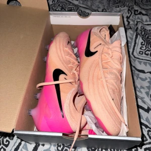Nike Phantom rosa fotbollsskor - Säljer ett par Nike Phantom fotbollsskor i ljusrosa med svarta detaljer och rosa sula. Skorna har snörning och rund tå, samt en platt sula med dobbar för bra grepp på planen. Perfekta för dig som vill sticka ut på fotbollsplanen! Bara använt en gång! 