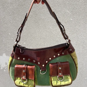 Grön och brun axelväska med detaljer - Säljer min ikoniska cowgirl väska från Osprey London i grönt och brunt med två fickor framtill, silvriga nitar och ringar. Väskan har en mix av äkta skinn (brun läder fram och bak samt axelrem), mocka (grön framsida) och faux ljusgrön krokodil skinn (baksida, fickor framtil). Har varit så förälskad i denna i flera år, men har köpt en annan nu så goodbye 🥲💚. OBS säljer billigare än vad den är värd då stora blixtlåset IBLAND stänger fel. Går att fixa!