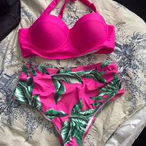 Rosa bikini med bladprint - Snygg bikini med vadderad rosa överdel och halterneckband. Höga trosor i matchande rosa med grönt bladprint. Perfekt för stranden eller poolen. Materialet känns mjukt och stretchigt.helt ny 