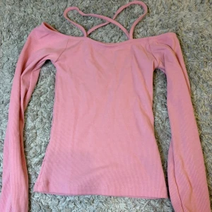 Rosa ribbad topp med smala axelband - Säljer en ljusrosa ribbad topp med långa ärmar och smala axelband som korsas i ryggen. Toppen har en offshoulder-design och är figurnära. Perfekt för vår och sommar!