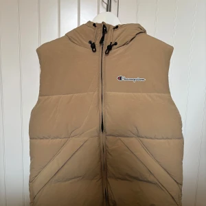 Beige dunväst från Champion med huva - Säljer en beige dunväst från Champion med huva och dragkedja framtill. Endast använd en gång, inga spår på användning.