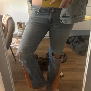 Unika jeans med orange paneler och slitningar - Säljer ett par ljusblå jeans med orange tygpaneler längs utsidan av benen. Byxorna har slitningar vid knäna och råa kanter. De har en normal passform och är bootcut . Perfekta för dig som vill sticka ut med en cool detalj!
