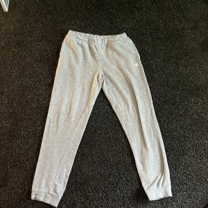 Grå mjukisbyxor från Champion - Säljer ett par grå mjukisbyxor från Champion med resår i midjan och vid bensluten. Byxorna har en liten Champion-logga på ena benet och är tillverkade i mjukt bomullsmaterial. Perfekta för en avslappnad stil.