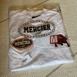 Vit t-shirt med patchar från Mercier - Säljer en oanvänd vit t-shirt från Mercier med lappen och förpackning kvar, med flera broderade patchar i rött, svart och vitt på bröstet och ärmarna. Stor Mercier-logga på ryggen. Klassisk passform och rund hals. Perfekt för dig som gillar streetwear och detaljer.