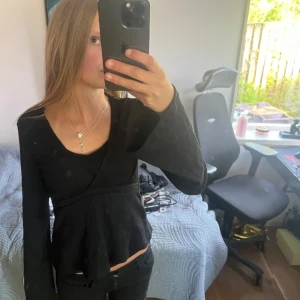 Svart omlottopp med knytband - Säljer en svart omlottopp med långa ärmar och knytband i midjan. Toppen har en v-ringad halsringning och är tillverkad i ett lätt, ribbat material. Perfekt för en stilren och trendig look. Man kan välja att ha linne under som jag har på bilden eller tröjan bara som den är🥰 endast avvänd 1 gång!