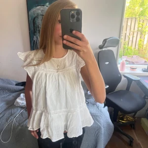 Vit volangblus med kort ärm - Säljer en söt vit blus med volangdetaljer och korta ärmar. Blusen har en lös passform och fina sömmar som ger extra struktur. Perfekt till sommaren och enkel att matcha med olika underdelar.