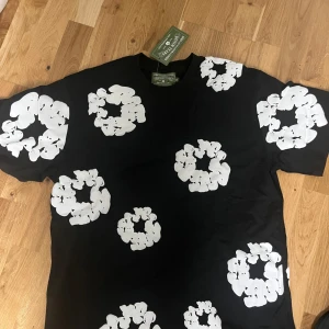 Svart t-shirt med vita blommor från Denim Tears - Säljer en svart t-shirt från Denim Tears med stora vita blommönster över hela tröjan. T-shirten har klassisk passform och är kortärmad. Perfekt för dig som vill sticka ut med en unik och trendig look.