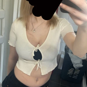 Vit croppad ribbad topp med knyt - Säljer en vit croppad topp med ribbad struktur och knyt framtill. Toppen har korta ärmar och djup v-ringning, perfekt för varmare dagar. Enkel och stilren design som passar till mycket.