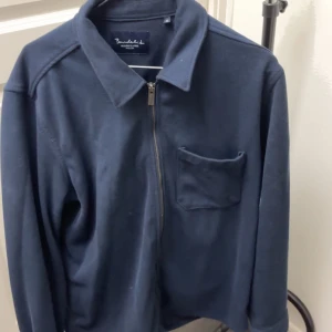 Mörkblå zip-jacka från Brandalised - Säljer en mörkblå jacka från Brandalised med dragkedja framtill och en bröstficka. Jackan har klassisk krage och lång ärm, perfekt för lager-på-lager. Enkel och stilren design som funkar till det mesta.