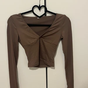 Brun croppad långärmad topp med v-ringning - Säljer en brun croppad topp med lång ärm och snygg v-ringning. Toppen har en figurnära passform och är perfekt att styla till jeans eller kjol. Enkel och stilren design som passar till många tillfällen.