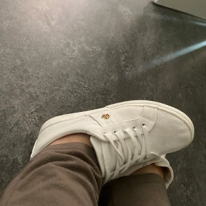 Vita sneakers från Lauren Ralph Lauren - Säljer ett par stilrena vita sneakers från Lauren Ralph Lauren med RL-detalj i guld på sidan. Skorna har klassisk snörning och är tillverkade i textilmaterial med en platt sula. Perfekta för en clean och fräsch look. Passar 39 med