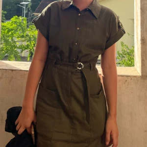 Olivgrön skjortklänning med bälte - Snygg olivgrön skjortklänning med korta ärmar och krage. Klänningen har knappar framtill, fickor och ett matchande bälte i midjan som ger en markerad siluett. Perfekt för en avslappnad men stilren look.