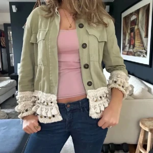 Ljusgrön jacka med virkade detaljer - Säljer en ljusgrön jacka med stora knappar och unika virkade volangdetaljer längst ner på ärmar och midja. Jackan har två bröstfickor och en croppad passform. Perfekt för dig som vill sticka ut!