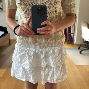 Vit volangkjol från KappAhl - Superfin vit kortkjol från KappAhl med bred smock i midjan och dubbla volanger. Kjolen är lätt och luftig, perfekt för varma dagar. Tillverkad i bomull med broderade detaljer för en söt look.