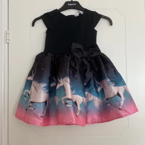 Svart klänning med enhörningar och rosett från HM - Superfin svart klänning med färgglad kjol med enhörningsmotiv och stor rosett i midjan. Kjolen har en snygg övergång från svart till rosa och blått, och klänningen har dragkedja i ryggen. Perfekt för dig som älskar magiska detaljer!