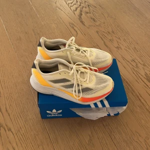Adidas löparskor med färgglada detaljer - Snygga Adidas löparskor i beige med grå ränder och färgglada detaljer i orange och gul. Skorna har en lätt och luftig design med snörning och en bekväm sula för optimal dämpning.