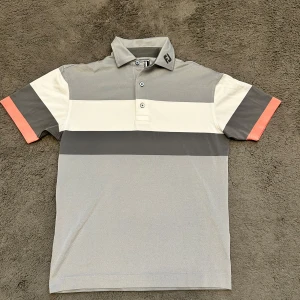 Grå och vit randig pikétröja från FootJoy - Snygg pikétröja från FootJoy i grått och vitt med breda ränder och en touch av orange på ärmarna. Tröjan har en klassisk krage och knappar framtill. Perfekt för en stilren look.