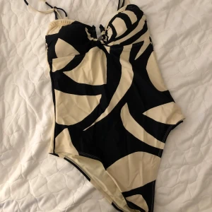 Mönstrad baddräkt från Second Female - Snygg baddräkt från Second Female i svart och beige mönster. Den har smala axelband och en dekorativ snörning framtill. Strl S, knappt använd