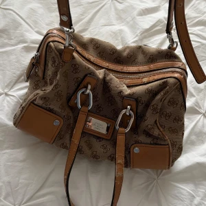 Beige och brun axelväska med monogram - Snygg beige axelväska med brunt monogrammönster och detaljer i brunt skinn. Väskan har flera dragkedjor och en justerbar axelrem. Perfekt för att bära dina dagliga nödvändigheter med stil.