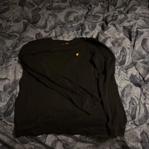 Svart långärmad tröja med gul logga - Säljer en stilren svart långärmad tröja från Lyle & Scott med en liten gul logga på bröstet. Perfekt för en avslappnad look. Passar bra till både jeans och chinos.