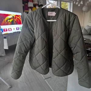 Olivgrön quiltad jacka från Nelly - Säljer en olivgrön quiltad jacka från Nelly. Jackan har en rund halsringning och stängs med knappar framtill. Perfekt för höst och vår med sin stilrena design och bekväma passform.