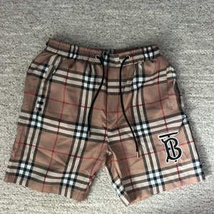 Rutig shorts med logotyp - Snygga rutiga shorts i beige, svart och röd med en framträdande logotyp på benet. De har en elastisk midja med snörning för justerbar passform. Perfekta för en avslappnad stil. Shorts/badbyxor