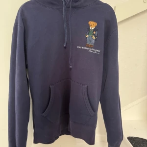 Mörkblå hoodie från Ralph Lauren - Säljer en snygg mörkblå hoodie från Ralph Lauren med en broderad teddy på framsidan. Hoodien är i bra skick inga tecken på utsidan från användning endast lite ludd inuti hoodien.