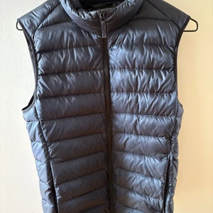  dunväst från Uniqlo i färgen navy blue - Säljer en stilren och lätt dunväst från Uniqlo. Västen har en dragkedja framtill och är perfekt för lager-på-lager under kyliga dagar. Den är designad för att hålla dig varm utan att kännas tung.