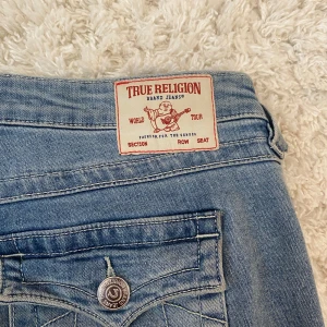 Blå jeans från True Religion - Säljer ett par snygga blå jeans från True Religion. De har en klassisk femficksdesign med märkets ikoniska logga på bakfickan. Jeansen har en bootcut-stil och är lågmidjade, vilket ger en avslappnad och trendig look. Strl 28