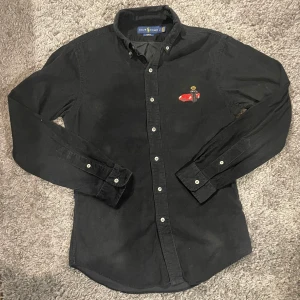 Svart skjorta från Ralph Lauren - Snygg svart skjorta från Ralph Lauren med broderad detalj av en röd bil och en figur på bröstet. Skjortan har en slim fit passform och långa ärmar med knappar. Perfekt för en stilren look.