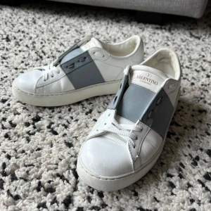 Valentino Open sneakers i vitt och grått - Snygga Valentino Open sneakers i vitt med gråa detaljer i stl 36,5. Skorna har en stilren design med helt nytt snöre och de ikoniska nitarna på hälen. Skorna är i bra skick, men har tecken på användning, se detaljer på bilderna. Hör av dig om du är intresserad eller vill ha fler bilder!