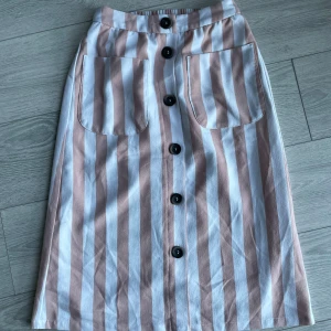 Randig kjol från ONLY - Säljer en snygg randig kjol från ONLY i beige och vit. Kjolen har stora fickor framtill och knäpps med svarta knappar längs hela framsidan. Perfekt för en stilren look!